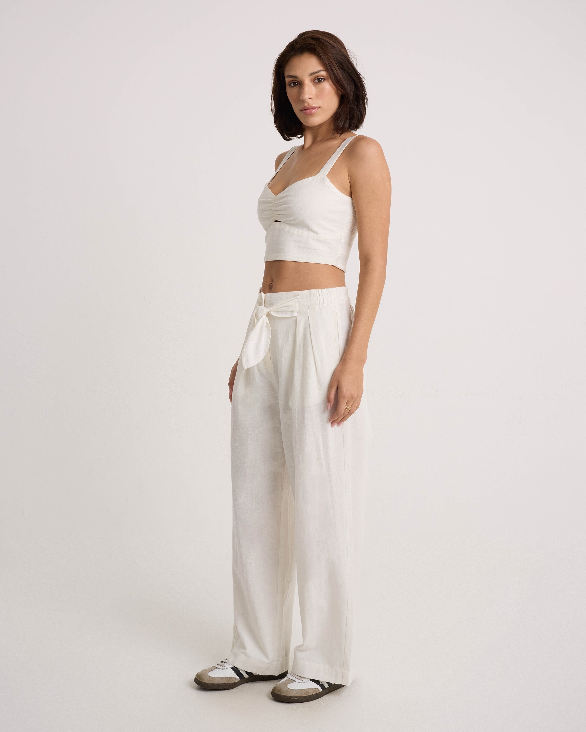 A woman with short dark hair poses against a plain light background, wearing Volcom's Women's Bow Chicka Pants—a high-waisted, wide-leg cotton-linen blend style with a tie-front waist—paired with a white crop top and white sneakers.