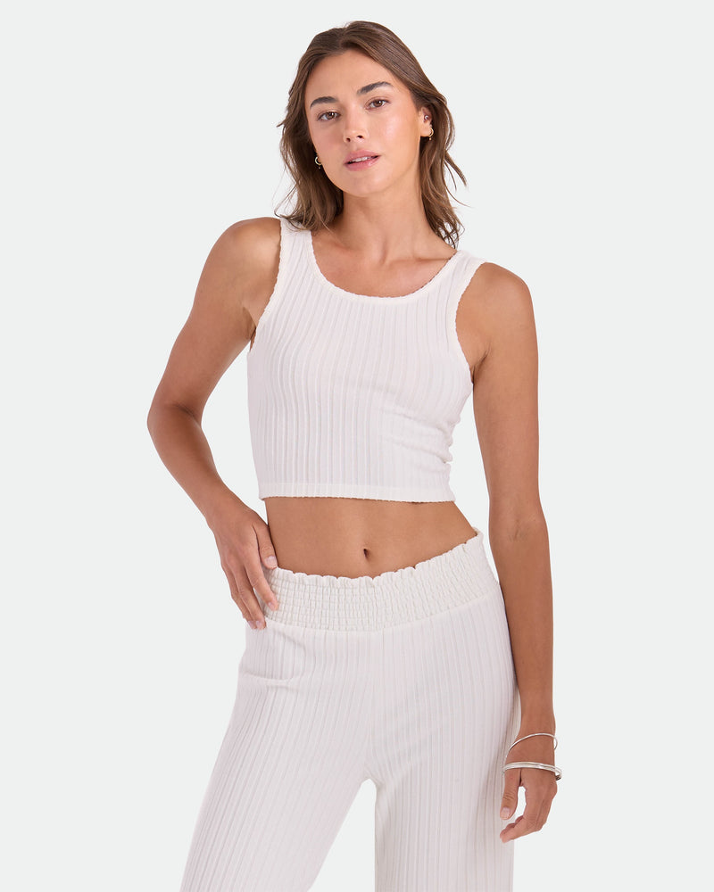 A woman poses against a light gray background wearing the Volcom Women's Lounge Bra Tank—a white, fitted scoop-neck style—paired with matching high-waisted pants. One hand rests on her hip while the other hangs by her side.
