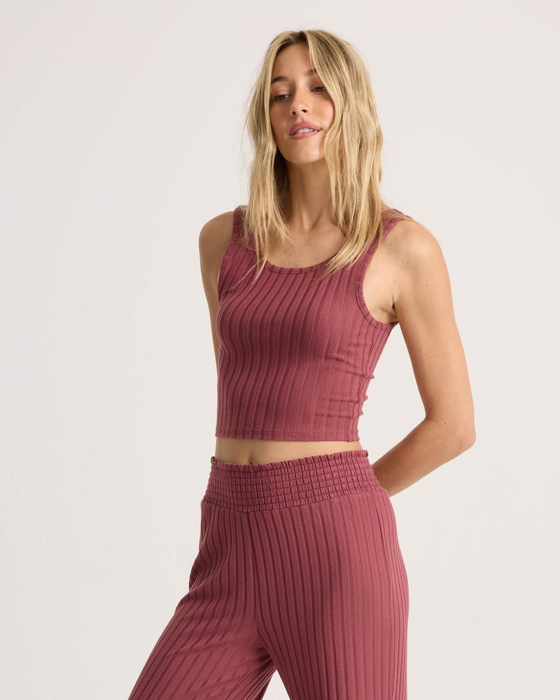 Model wearing the Volcom Women’s Sun Lounge Bra Tank, a fitted, sleeveless, ribbed crop top in rose, paired with matching high-waisted pants. This versatile knit ensemble combines comfort and style, perfect for any occasion.