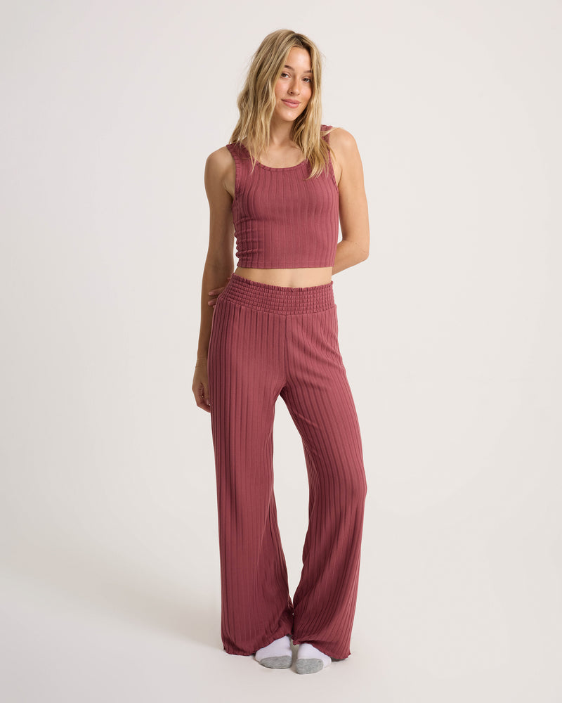 A woman stands against a plain background wearing the Volcom Women’s Sun Lounge Bra Tank, a fitted scoop-neck, sleeveless, ribbed, rose-colored crop top paired with matching wide-leg pants. She is barefoot and smiling slightly.