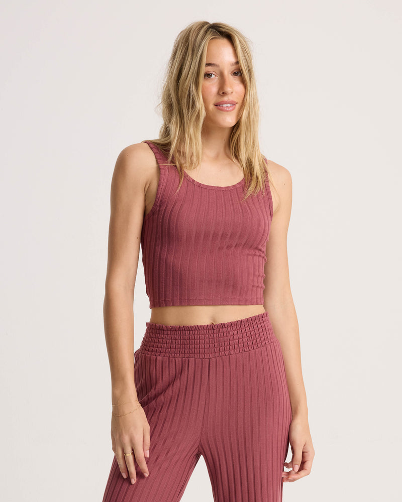 A woman with long blonde hair wears the Volcom Women’s Sun Lounge Bra Tank—a fitted, scoop-neck, ribbed mauve crop top—paired with matching high-waisted pants, standing against a plain light background.