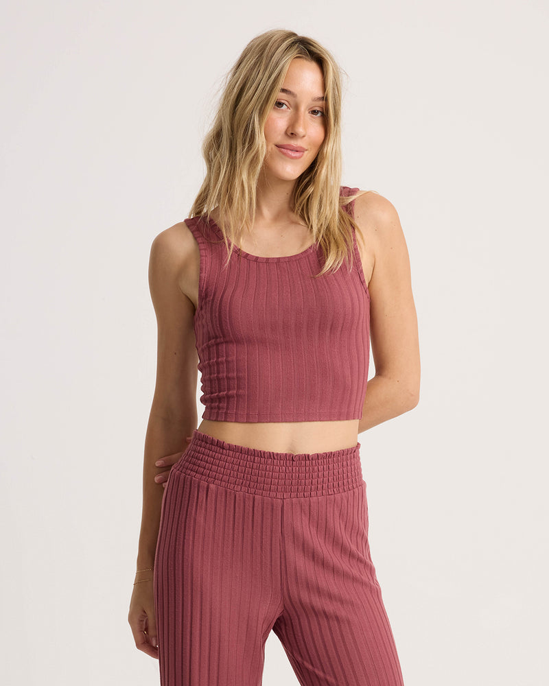A woman stands against a plain white background, smiling softly at the camera while wearing the Volcom Women’s Sun Lounge Bra Tank—a versatile, fitted mauve knit with a sleeveless scoop-neck—paired with matching high-waisted pants.