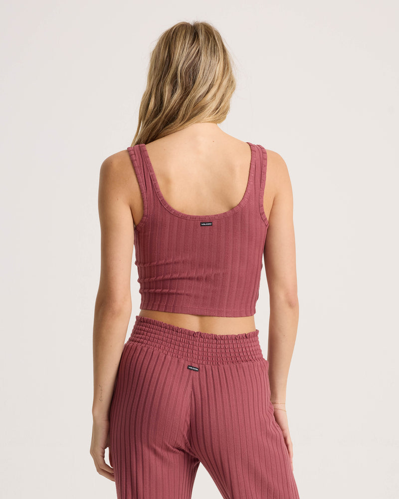 A person with long, wavy blonde hair is seen from the back against a plain light background, wearing the Volcom Women’s Sun Lounge Bra Tank—a matching ribbed, mauve-colored fitted scoop-neck crop tank top—paired with high-waisted pants.