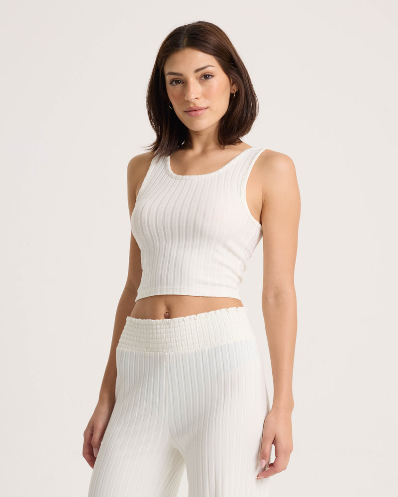 A woman with shoulder-length brown hair wears the Volcom Women’s Lounge Bra Tank—a white, ribbed, fitted scoop-neck top—paired with matching high-waisted pants, standing against a plain light background.