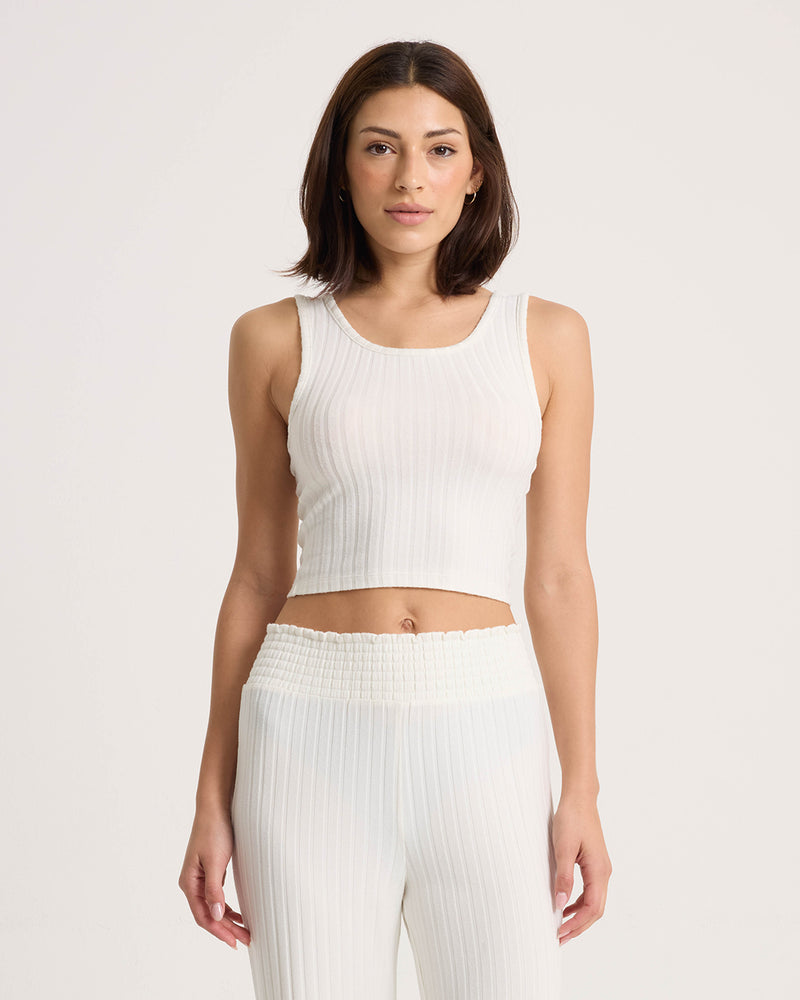 A woman with shoulder-length brown hair stands against a plain light background, wearing the Volcom Women’s Lounge Bra Tank—a fitted, sleeveless white ribbed crop top—paired with matching high-waisted pants.