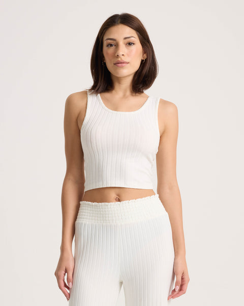 A woman wearing the Volcom Women’s Lounge Bra Tank—a fitted, scoop-neck, ribbed white crop top—paired with matching high-waisted pants stands against a plain light background, looking at the camera.