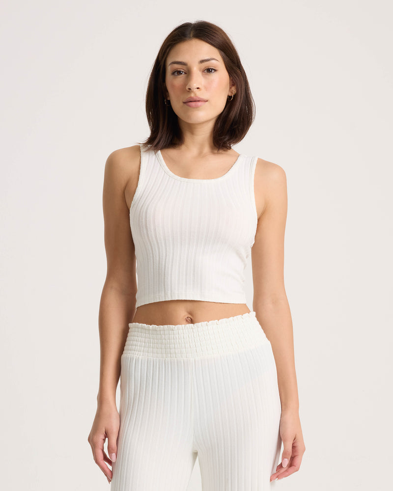 A woman wearing the Volcom Women’s Lounge Bra Tank—a fitted, scoop-neck, ribbed white crop top—paired with matching high-waisted pants stands against a plain light background, looking at the camera.