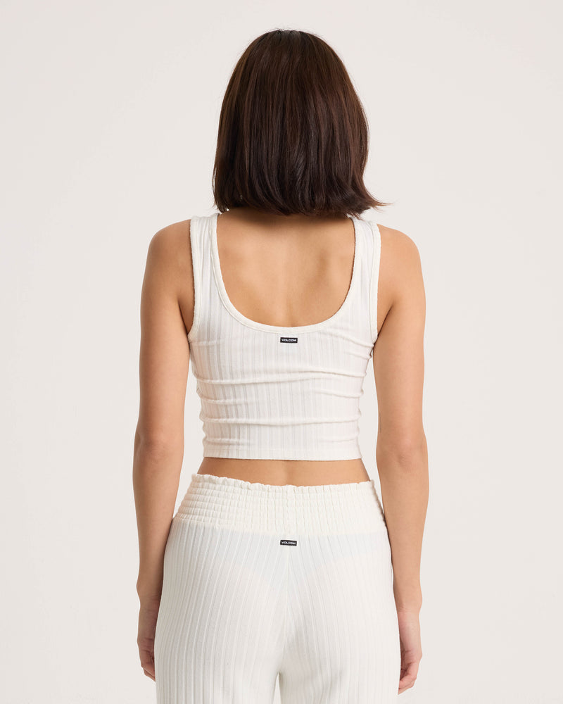 A woman with shoulder-length brown hair is shown from the back, wearing the Volcom Women’s Lounge Bra Tank—a fitted scoop-neck white ribbed top—paired with matching white ribbed pants against a plain light background for an everyday essential look.
