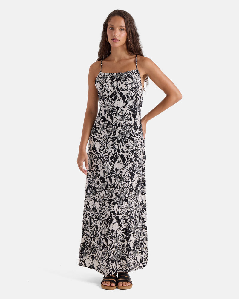 A woman models the Volcom Women's Slip Maxi, featuring a black and white tropical leaf pattern, fluid silhouette, and thin straps. She pairs it with black sandals against a plain white background.