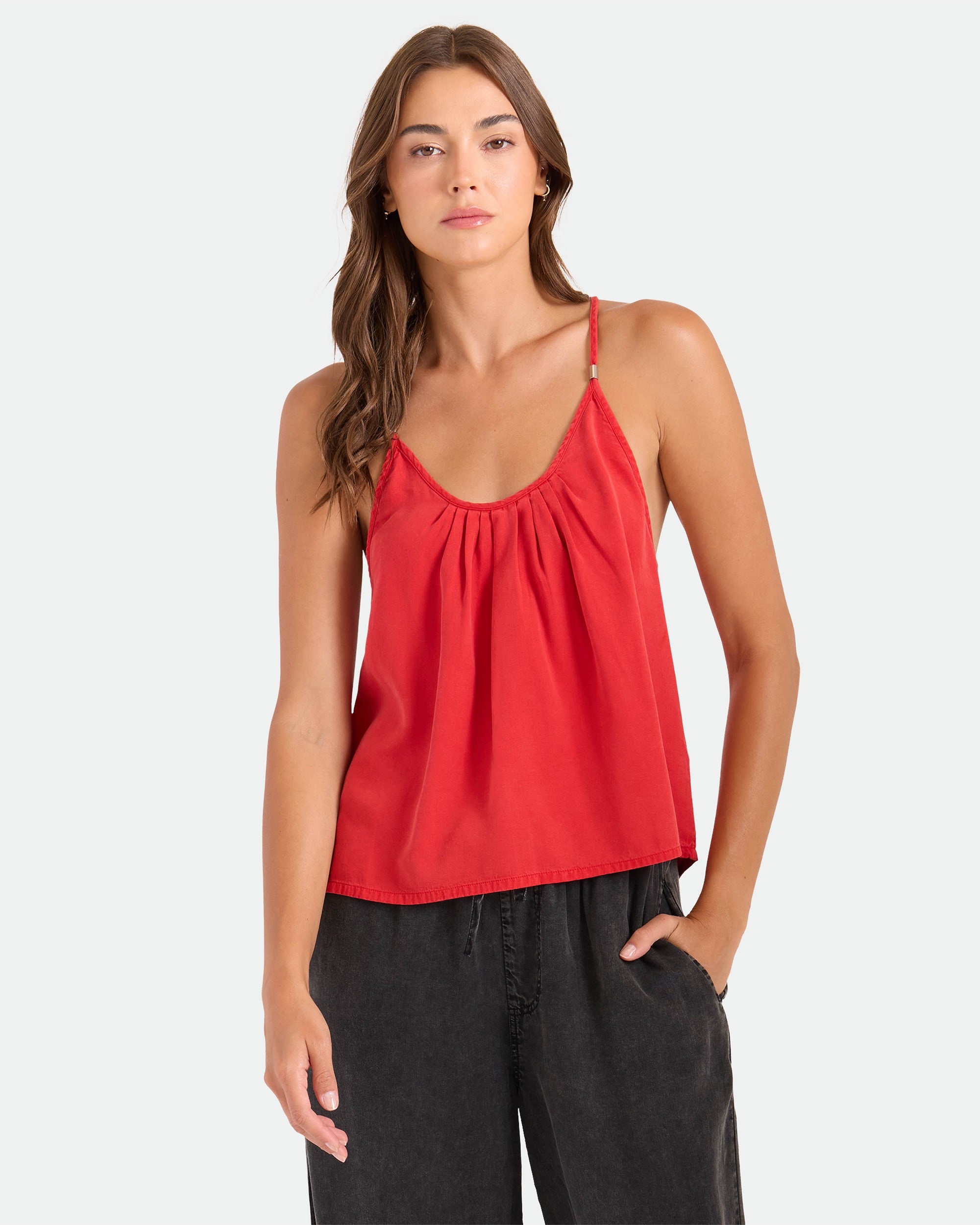 A woman stands against a light backdrop, looking at the camera with a neutral expression, wearing Volcom's Women's Pleat Heart Cami in persimmon and relaxed fit dark pants. One hand is in her pocket.