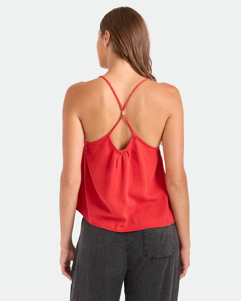 A woman with brown hair worn down faces away, wearing the Volcom Women's Pleat Heart Cami in Persimmon with crisscross straps and a relaxed fit, paired with dark gray pants against a plain white background.