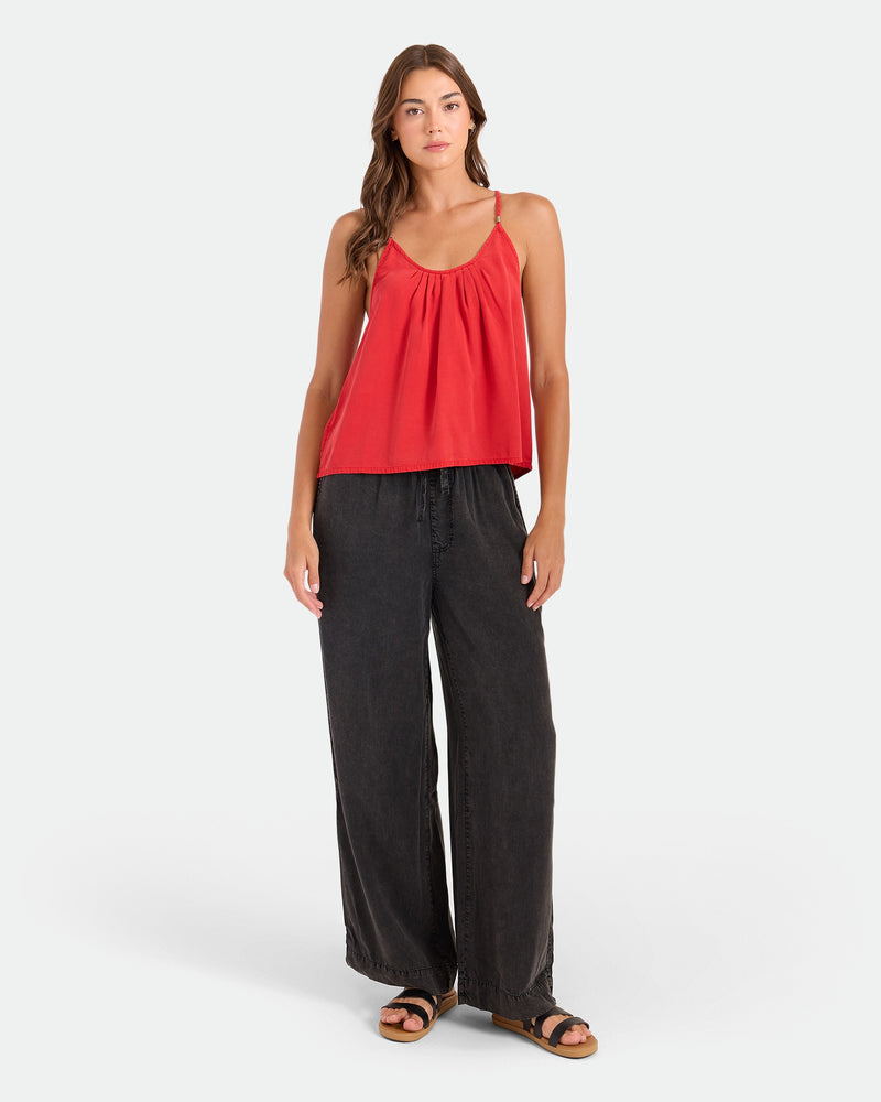 A woman with long hair wears the Volcom Women's Pleat Heart Cami in persimmon, paired with relaxed wide-leg black pants and black sandals, standing casually against a plain background.
