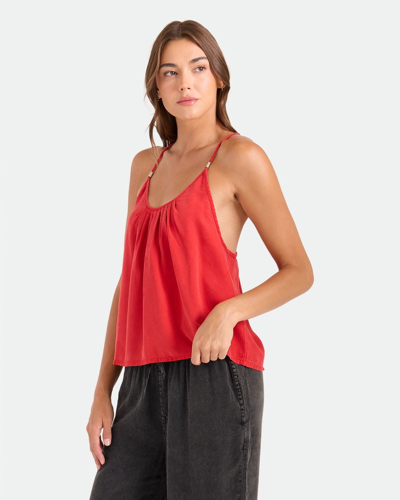 A woman in a relaxed fit Volcom Women's Pleat Heart Cami in persimmon and dark gray pants stands against a plain light background, gazing slightly left with a neutral expression.