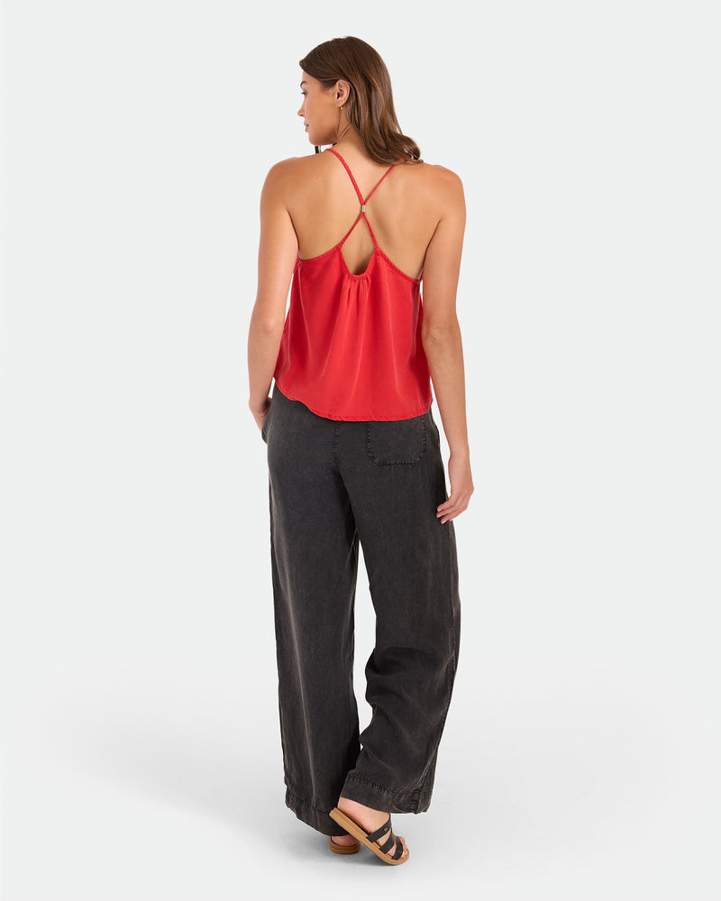 A woman with brown hair, wearing the Volcom Women's Pleat Heart Cami and loose black pants, stands facing away in sandals against a plain white background.
