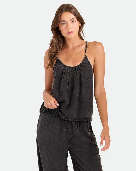 A woman with long brown hair wears a loose-fitting, dark Women's Pleat Heart Cami by Volcom and matching drawstring pants, standing against a plain light background.