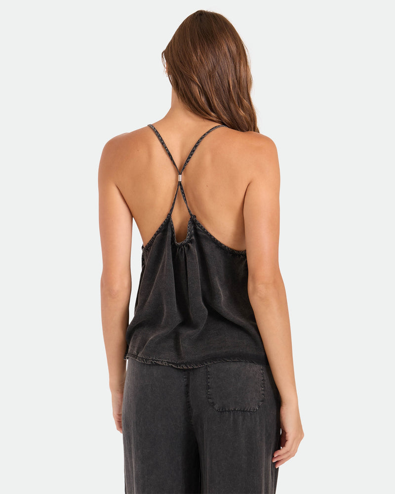 A woman seen from behind wears the Volcom Women's Pleat Heart Cami—a black, loose-fitting, strappy tank with an open back and matching black pants, featuring a slightly faded, washed look.