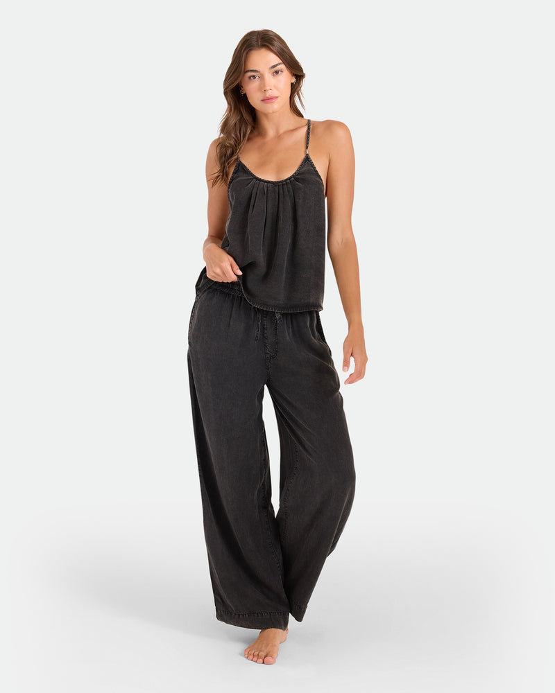 A woman stands barefoot in a Volcom Women's Pleat Heart Cami and matching wide-leg black pants against a plain white background.