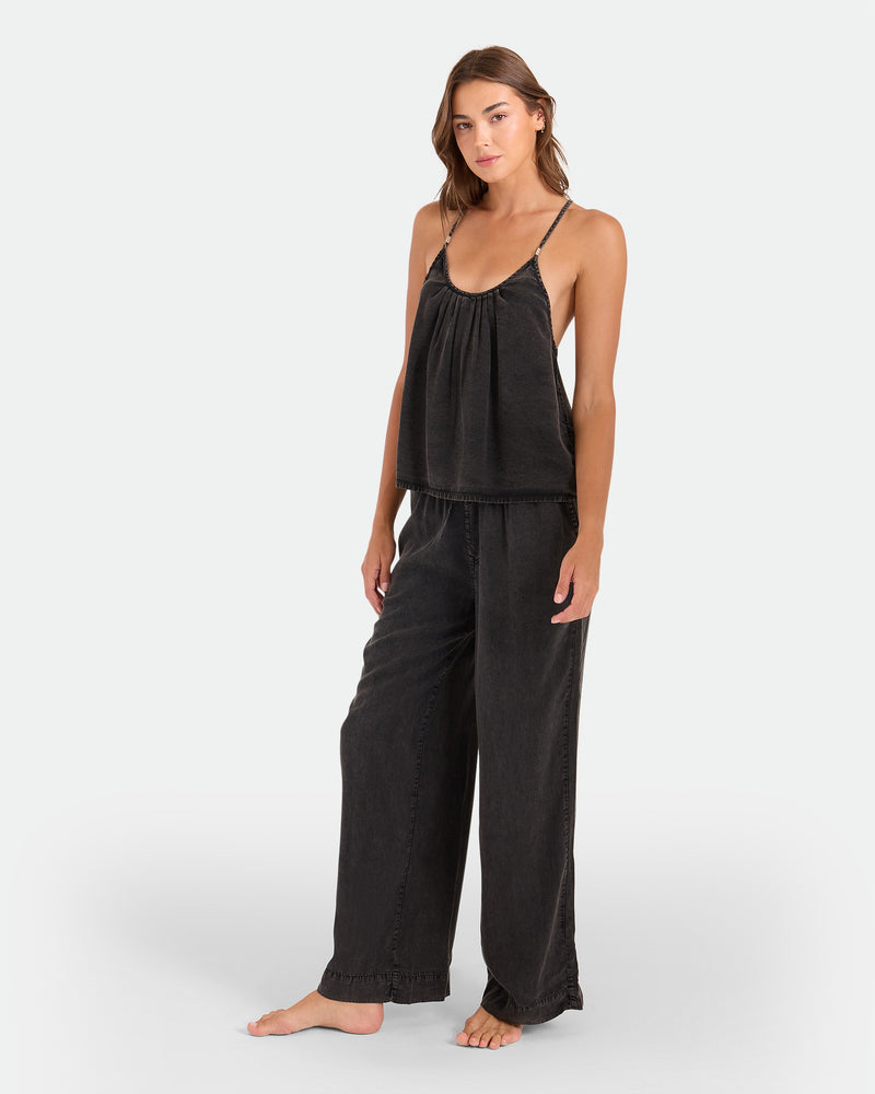 A woman stands barefoot in a loose-fitting, black Women's Pleat Heart Cami by Volcom with spaghetti straps and matching wide-leg pants against a plain white background.