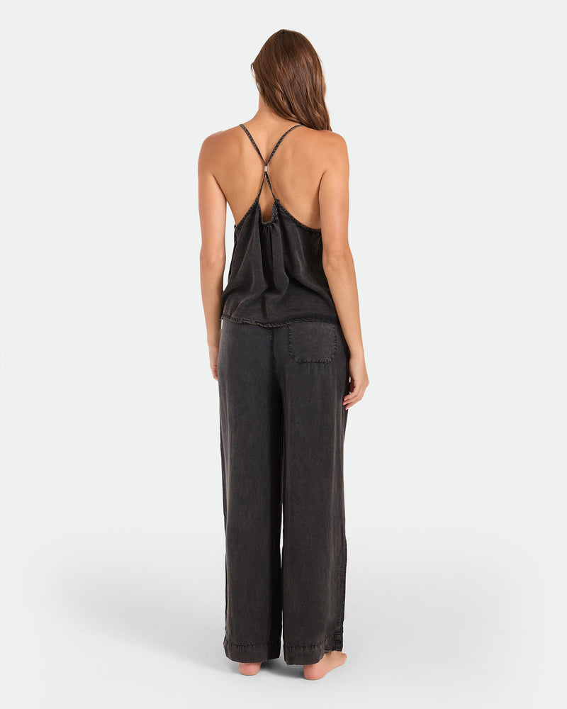A woman stands barefoot facing away, wearing the Volcom Women's Pleat Heart Cami—a loose-fitting, strappy black tank with crisscrossed straps—paired with matching wide-leg pants, set against a plain white background.
