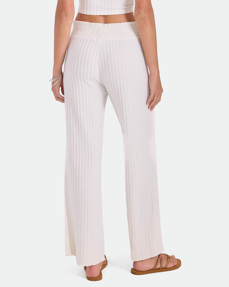 A person is seen from the back wearing Volcom Women's Sun Lounge Pants—high-waisted, wide-leg white pants with a relaxed fit and elastic waistband—paired with brown sandals and a cropped, sleeveless top for an effortlessly comfortable look.