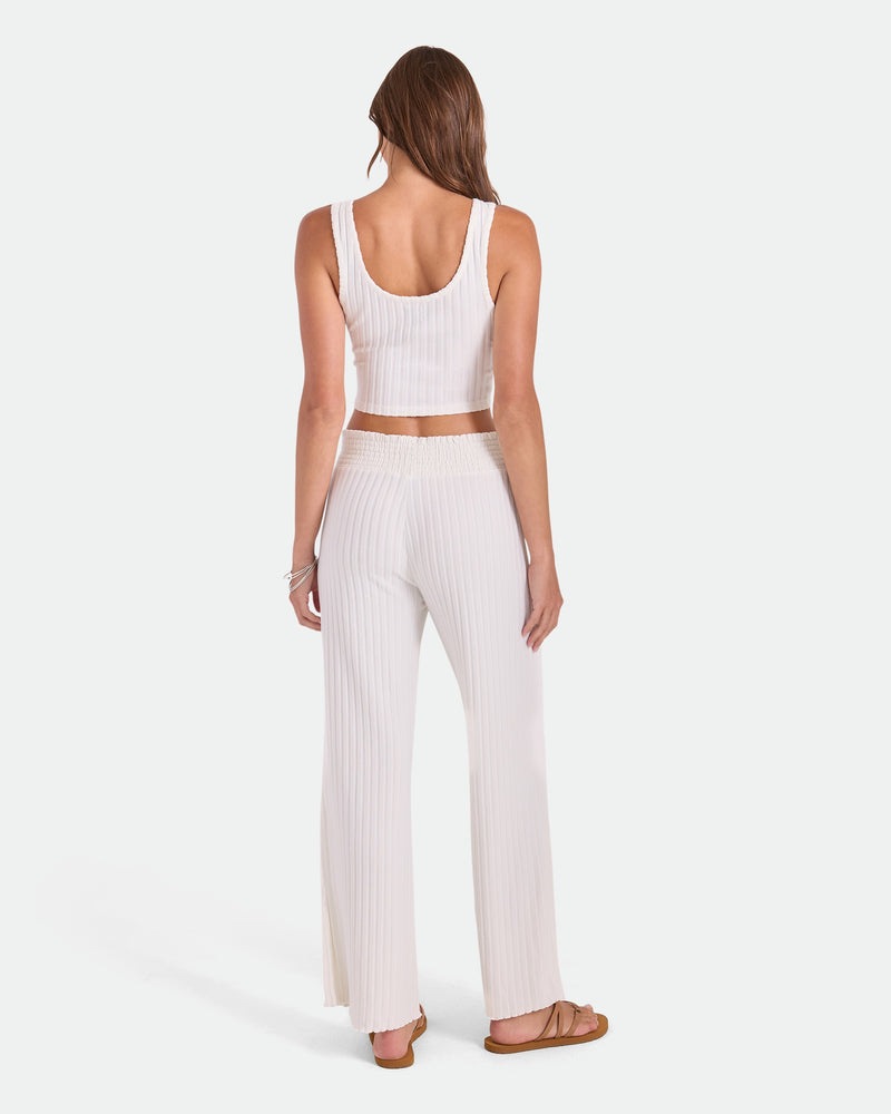 A woman with long brown hair faces away, wearing Volcom Women's Sun Lounge Pants—relaxed fit with elastic waistband—and a white ribbed crop top and sandals, shown on a plain white background.