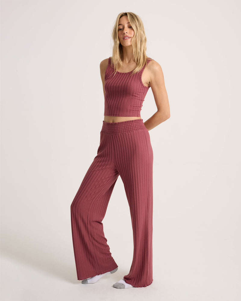A person wearing the Volcom Women’s Sun Lounge Pant in dusty rose, styled with a matching ribbed, sleeveless crop top, stands against a plain white background with hands behind their back and one foot slightly forward. The wide-leg lounge pant features an elasticated waistband for comfort.