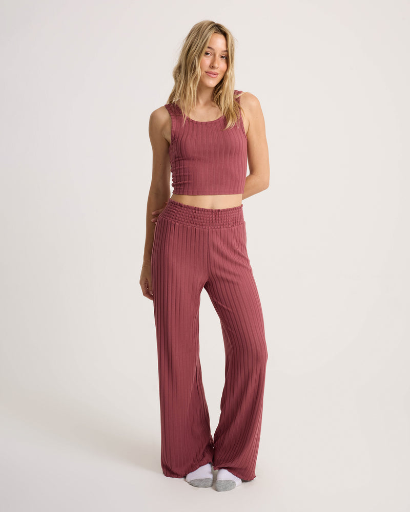 A woman stands against a plain light background, smiling slightly with one hand resting by her side, wearing the Volcom Women’s Sun Lounge Pant—a sleeveless, ribbed mauve-colored lounge set featuring wide-leg pants with an elasticated waistband.