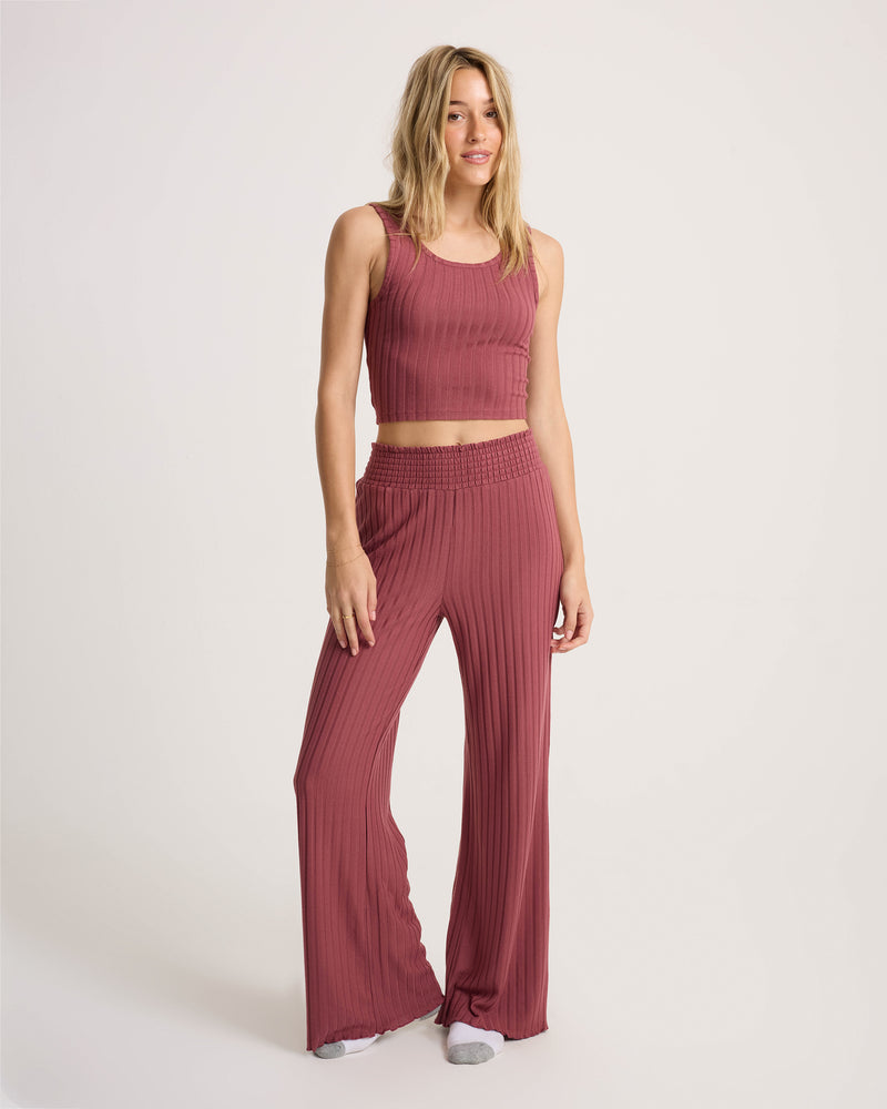 A woman wearing the Volcom Women’s Sun Lounge Pant, featuring a high-waisted fit with an elasticated waistband, pairs it with a sleeveless, ribbed mauve crop top as she stands against a plain white background, smiling with one leg slightly bent.