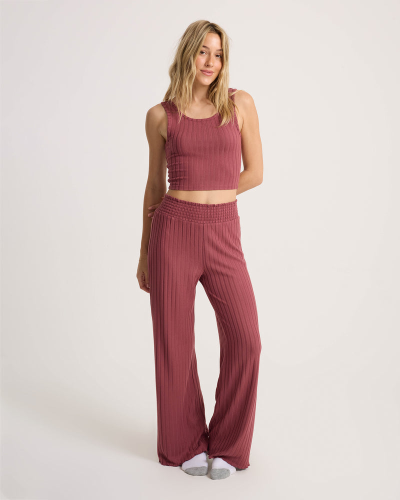 A person stands against a plain background wearing Volcom’s Women’s Sun Lounge Pant—a matching ribbed mauve, wide-leg pant with an elasticated waistband—paired with a sleeveless crop top; their left hand relaxed at their side and white socks visible.