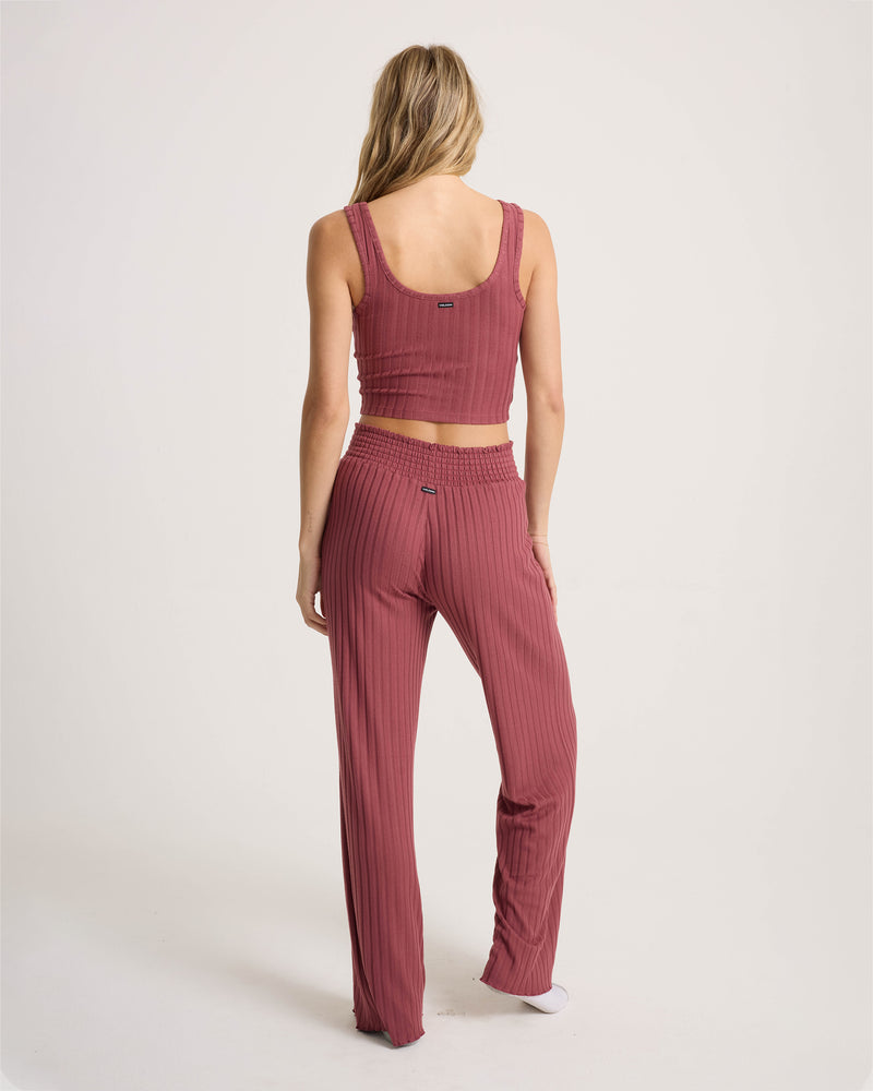 A woman with long blonde hair wears the Volcom Women’s Sun Lounge Pant, a mauve-colored, high-waisted lounge pant with an elasticated waistband, paired with a matching sleeveless ribbed crop top; she stands barefoot and faces away against a plain white background.