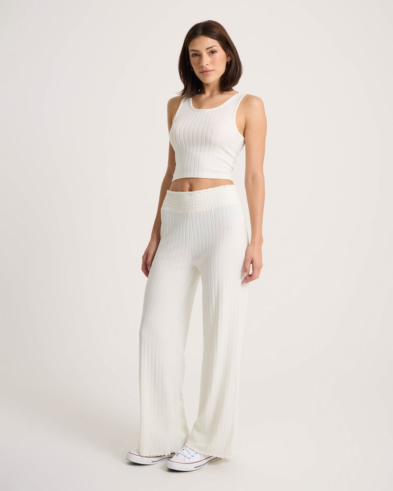 A woman stands against a plain background, wearing Volcom’s Women’s Sun Lounge Pants paired with a white ribbed sleeveless crop top and white sneakers. Her straight, shoulder-length hair and neutral expression complement the relaxed fit of her soft, stretchy knit outfit.