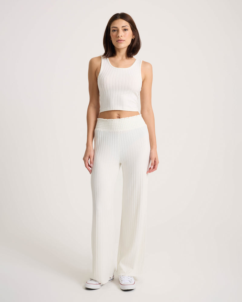 A woman stands against a plain background wearing the Volcom Women’s Sun Lounge Pants, featuring a smocked elastic waistband for comfort, paired with a sleeveless, ribbed white crop top and finished with white sneakers for a relaxed look.