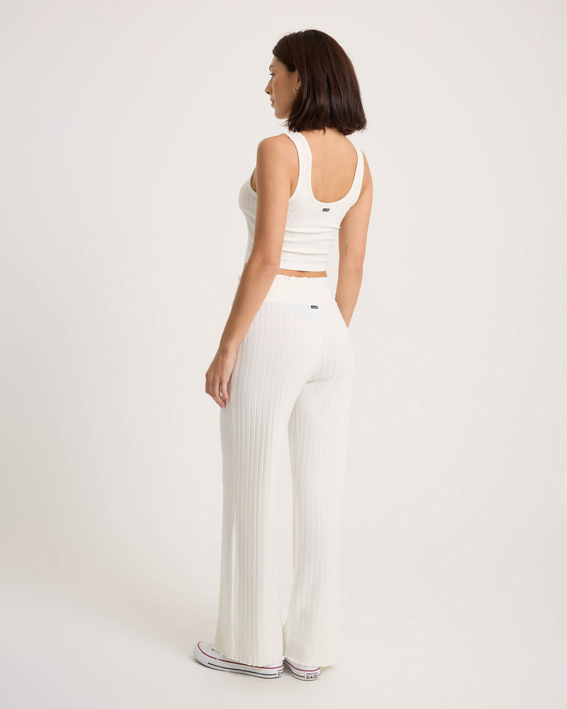 A woman with short dark hair is wearing a white sleeveless crop top and Volcom Women’s Sun Lounge Pants—high-waisted, ribbed pants featuring a relaxed fit and smocked elastic waistband—as she stands sideways, looking slightly away from the camera against a plain background.