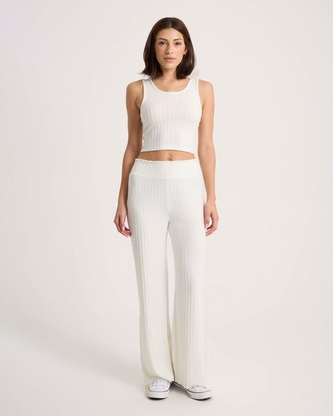 A woman stands against a plain light background, wearing a sleeveless white ribbed crop top and Volcom Women’s Sun Lounge Pants in a matching high-waisted relaxed fit, paired with white sneakers.