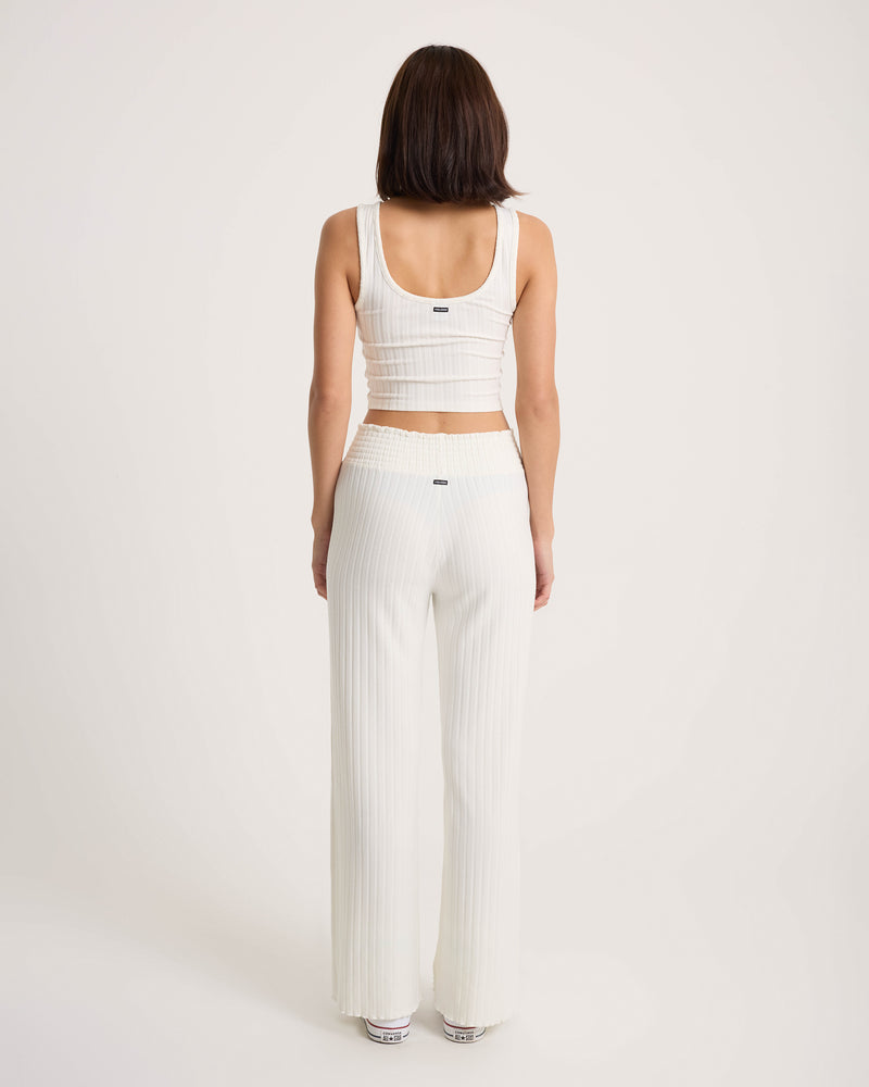 A woman with dark hair, seen from behind, stands against a plain light background wearing Volcom Women’s Sun Lounge Pants—a high-waisted, wide-leg lounge style with a smocked elastic waistband—paired with a sleeveless white ribbed crop top.