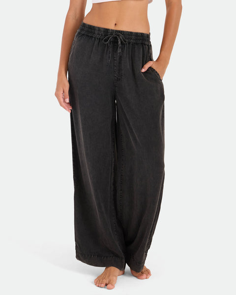 A person models Volcom's Women's Washed Up Twill Pants—loose-fitting, high-waisted black pants with an elastic waist and wide-leg cut—barefoot on a white background, hand in pocket to highlight their relaxed, casual style.