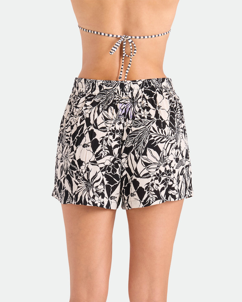 A woman is shown from the back wearing Volcom's Women's Funtastical Viscose Shorts with a black and white floral print and elastic waistband, paired with a striped string bikini top against a plain light background.