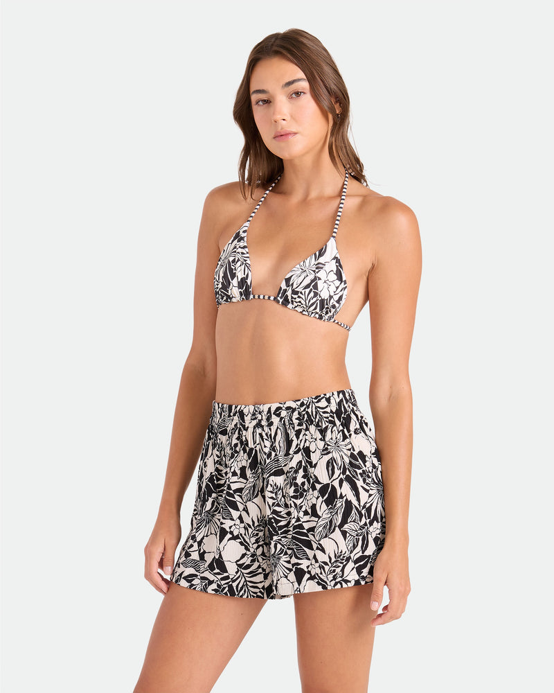 A woman wears Volcom's Women's Funtastical Viscose Shorts with a matching Black Ecru bikini top, featuring an elasticated waistband, as she stands against a plain light background, looking confidently at the camera.
