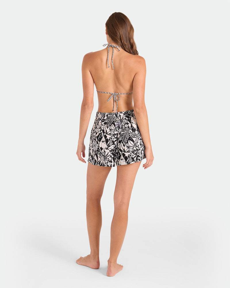 A woman faces away, barefoot against a white background, wearing Volcom Women's Funtastical Viscose Shorts with an elastic waistband in a Black Ecru print and a halter-neck striped bikini top.