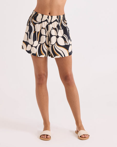 A model showcases the Volcom Women's Funtastical Shorts—high-waisted pull-on shorts featuring an elastic waist and a relaxed fit with an abstract black, white, orange, and beige print. Paired with beige sandals, the look is styled against a plain white background for a comfortable yet stylish ensemble.
