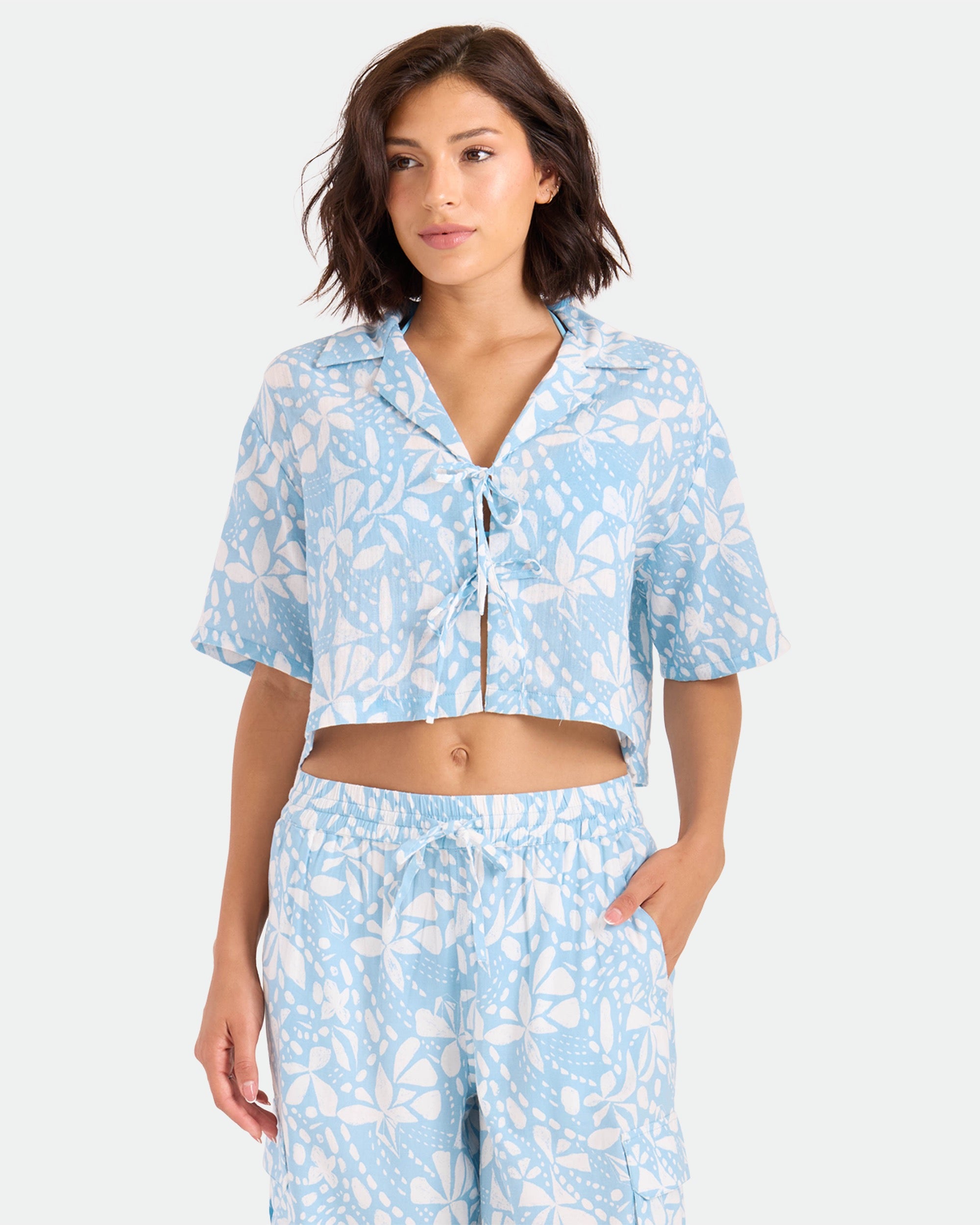 A woman in the Volcom Women's Tied Up Crop Shirt, featuring a light blue and white floral pattern, stands with one hand in her pocket against a plain light background.