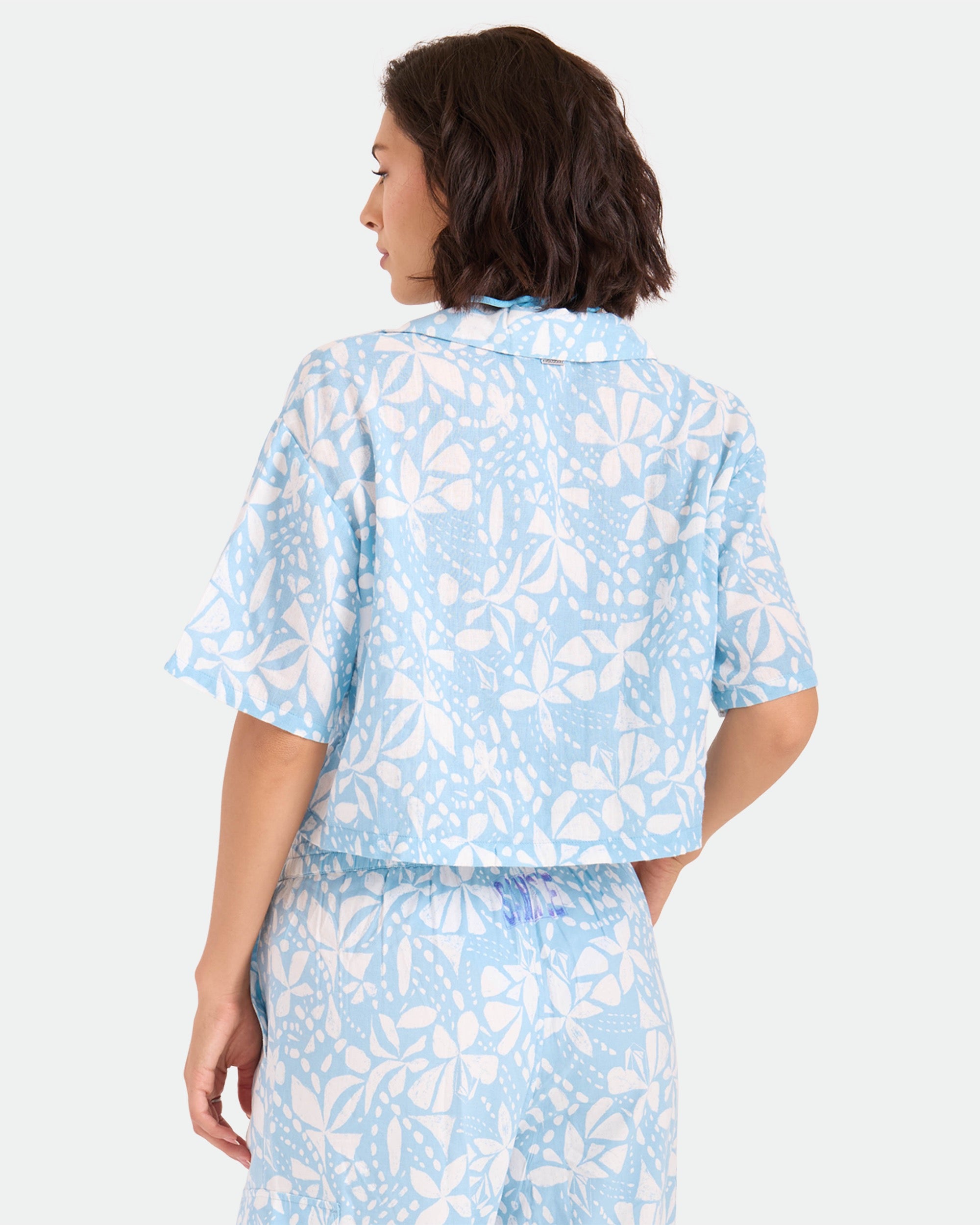 A woman with short dark hair is seen from the back, wearing Volcom's Women's Tied Up Crop Shirt and matching pants in a light blue and white pattern, set against a plain light gray background.