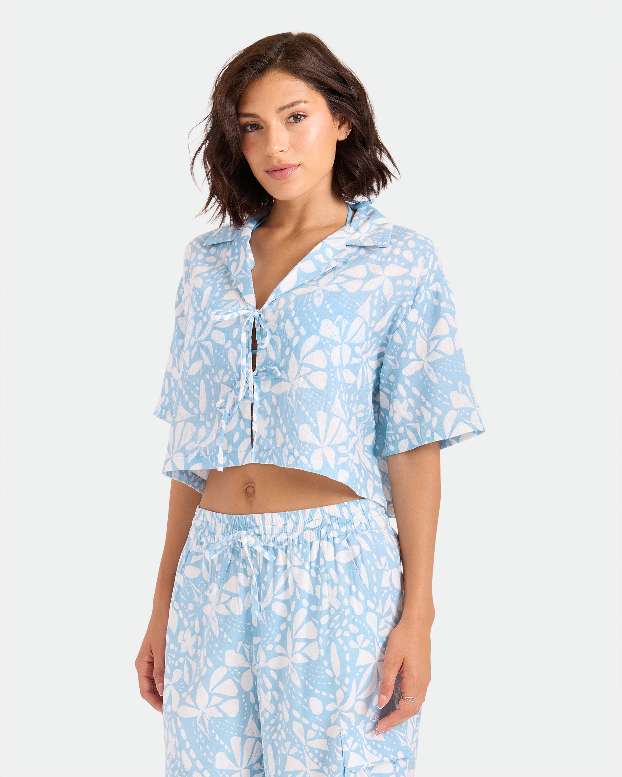 A woman models the Volcom Women's Tied Up Crop Shirt, a light blue and white floral cotton pajama set with a short-sleeve tie-front crop top and matching loose pants, standing against a plain light background.