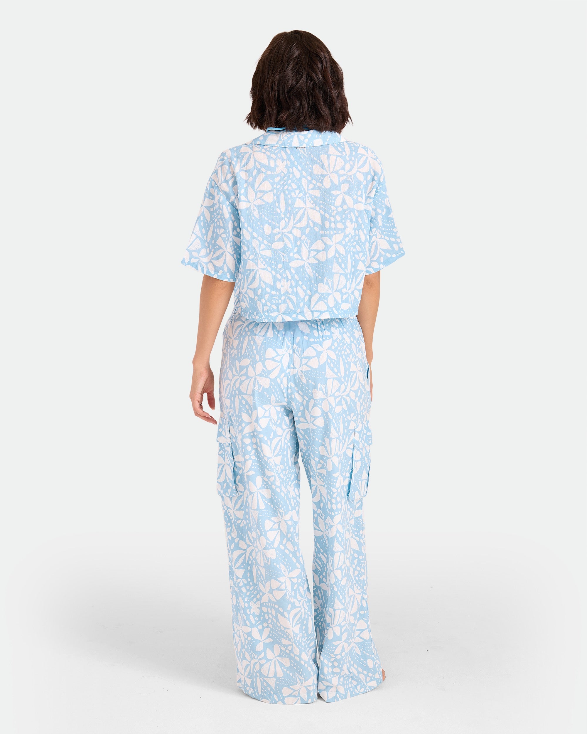 A woman with shoulder-length dark hair stands facing away, wearing the Volcom Women's Tied Up Crop Shirt in a light blue and white floral cotton pattern with matching wide-leg pants against a plain white background.