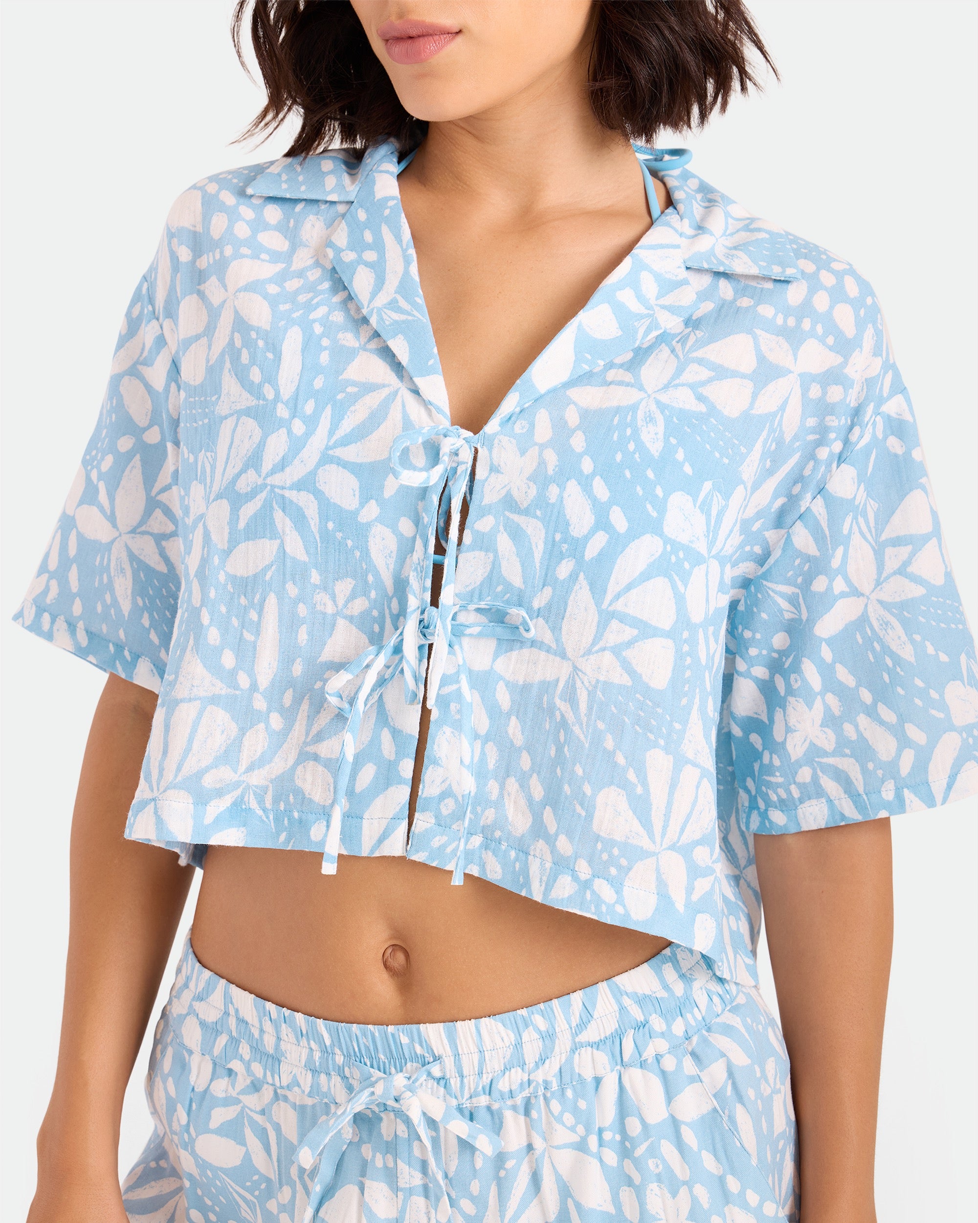 A woman wears the Volcom Women's Tied Up Crop Shirt—light blue and white, floral-patterned, short-sleeved with a tie front—paired with matching drawstring shorts for a casual, relaxed summer look.