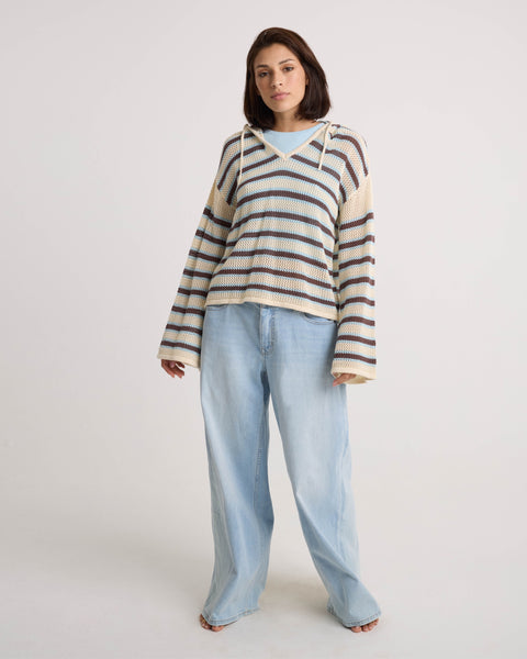 A woman with shoulder-length brown hair and a relaxed expression wears the Volcom Women's Bonfire Hoodie—a loose, cream-and-navy striped knit with crochet texture and wide sleeves—paired with light blue wide-leg jeans against a white background.