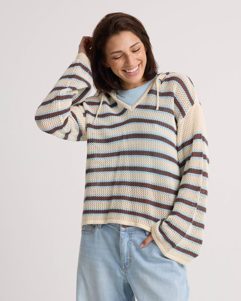 A smiling woman with short brown hair models the Volcom Women's Bonfire Hoodie—a loose, striped cotton sweater with a crochet texture—paired with light blue jeans. She poses against a plain background, hand in pocket and touching her head.