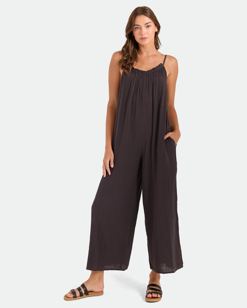 A woman with long hair wears the Volcom Women's Ruffle Up Romper, a loose, sleeveless, dark romper with spaghetti straps and wide legs. She stands with one hand in her pocket and wears black sandals against a plain white background.