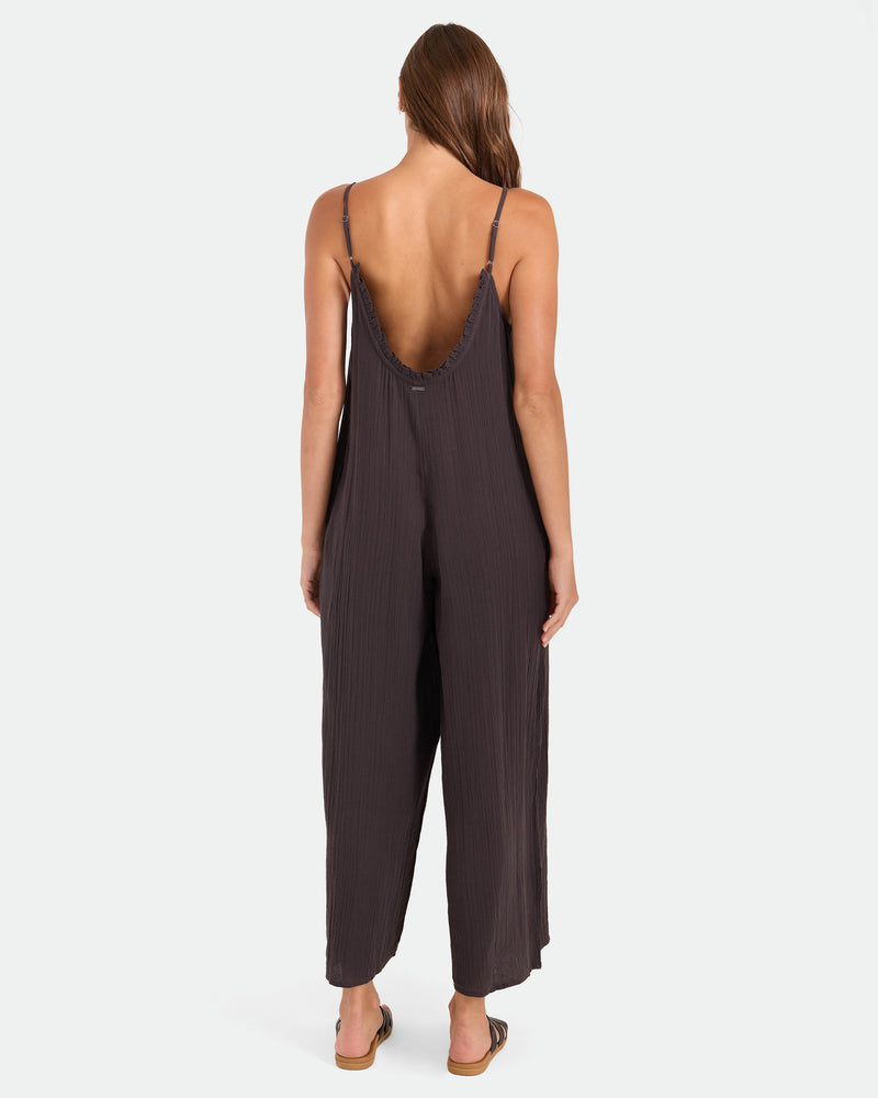 A woman with long brown hair, in flat sandals, wears the Volcom Women's Ruffle Up Romper—a loose, dark brown sleeveless jumpsuit with thin straps and a low back. She stands facing away against a plain white background.