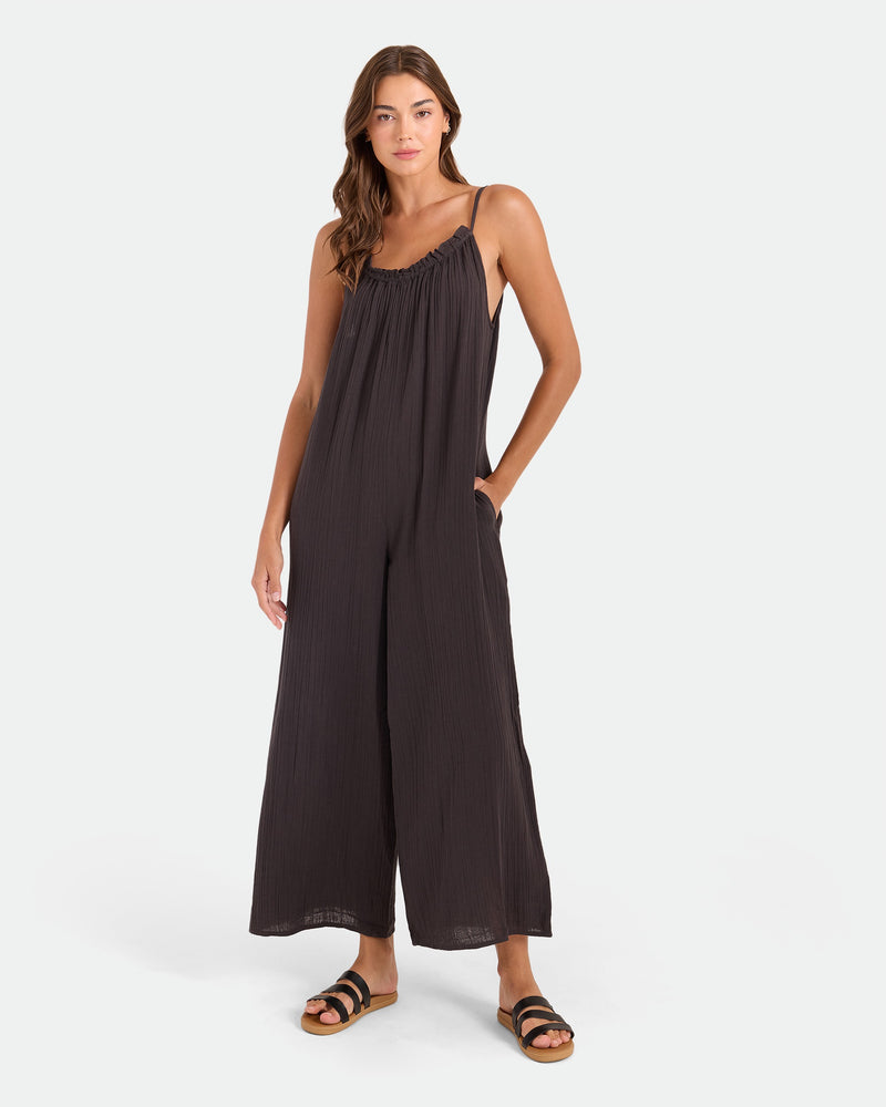 A woman poses against a plain background in the Volcom Women's Ruffle Up Romper—a loose, sleeveless black jumpsuit with thin straps and side pockets. She wears black flat sandals and has long, wavy hair.